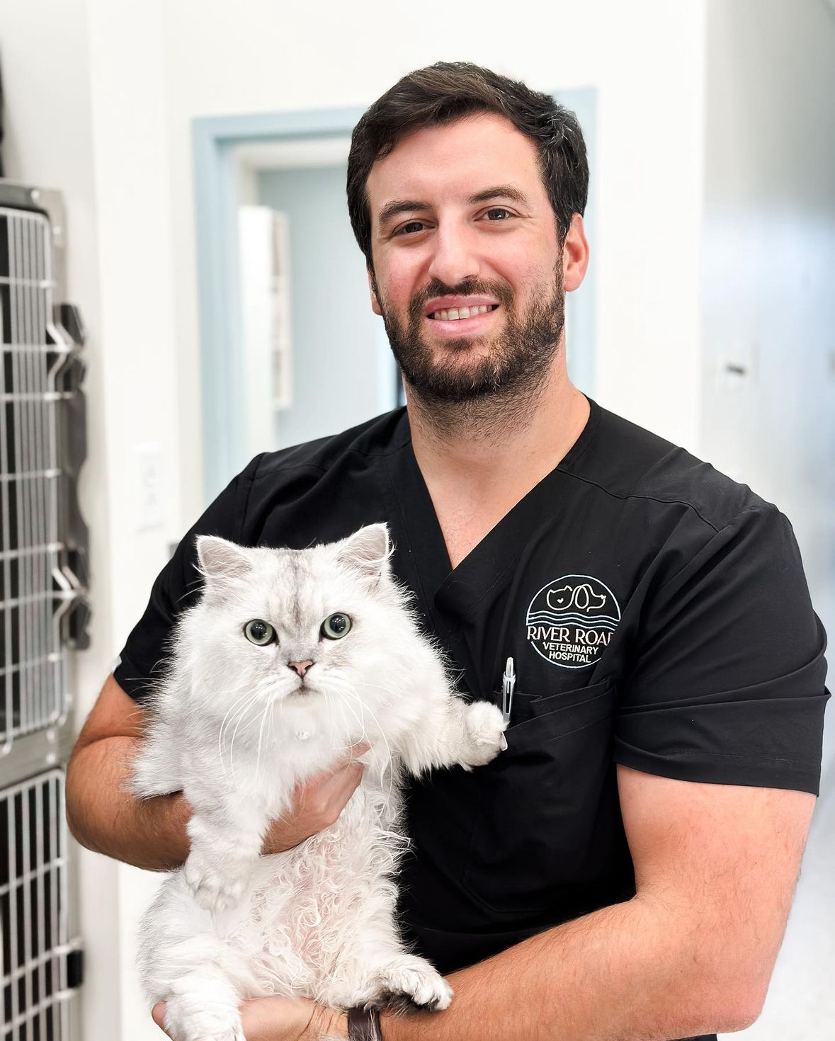 IMG_8146 A smiling man in black scrubs holds a fluffy white cat inside a veterinary clinic. The man has dark hair and a beard, and the cat has big green eyes. The clinic setting includes a silver cage and light-colored walls.