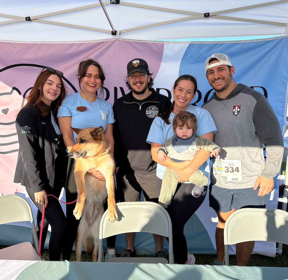 Screen Shot 2023-10-30 at 5.29.52 PM Five adults and a toddler gather around a dog under a tent, smiling for the photo at an outdoor event.
