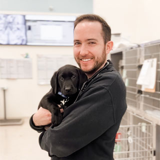 A man smiling at the camera, holding a black labrador puppy in his arms in a room that seems to be a veterinary clinic.